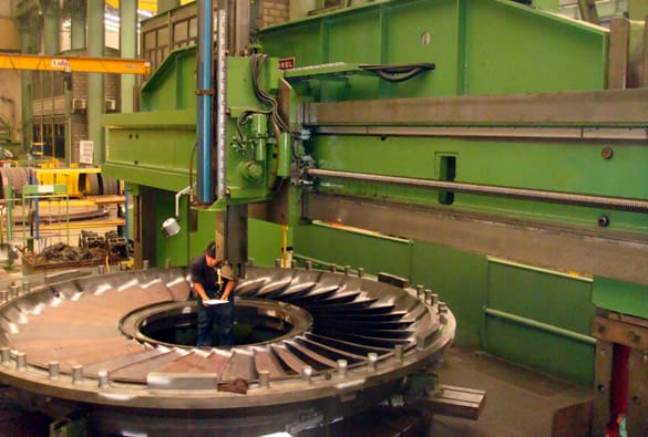An industrial machine operator working on a large, circular metal component with intricate blades, likely part of a turbine or heavy-duty machinery. The component is mounted on a massive green machine tool in a factory setting.