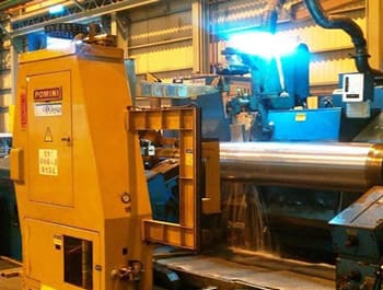 Industrial roll grinder in operation, with large metal rollers being processed. The machinery is designed for precision grinding.
