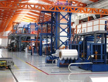Interior of a modern steel production facility with large rolls of processed metal in the foreground and industrial machinery in the background.